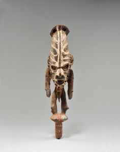 Bouchon de flûte sacrée, Biwat, rivière Yuat, région du bas Sepik. Ces figures wusear couronnaient les grandes flûtes sacrées en bambou haiyang par lesquelles la « voix » des ancêtres se faisait entendre lorsque l’instrument était joué. Possession d’un groupe héréditaire, elles étaient conservées dans les maisons des hommes, leur signification, à la fois sociale, cérémonielle et religieuse, étant considérable. Cet exemplaire est peint de lignes blanches semblables aux peintures faciales que l’on trouve à la jonction de la région Biwat et de la rivière Sepik. Dépourvu de sa parure en plumes de casoar et de ses ornements divers — croissant de nacre traversant la cloison nasale, dents de phacochère, chaînettes d’anneaux en écaille de tortue aux oreilles, coquillages, armature de sparterie fixant au menton une barbe de cheveux humains —, il représente un homme debout, sur un piquet. Ces figurations se caractérisent par une large tête dont la puissance s’exprime à travers la projection du crâne en pain de sucre, par un tronc grêle porté par des jambes puissantes, des bras ballants et un phallus pendant à hauteur des genoux. Les traits du visage sont concentrés sous un front surdimensionné avec des pommettes saillantes, un nez busqué puissant et une large bouche ouverte, aux lèvres charnues, une arcade sourcilière épaisse et des yeux incrustés de fragments de nacre pour figurer les pupilles. Bois et pigments. H. : 41,3 cm. Offert par la baronne de Béthune, en 1931, aux MRAH, en mémoire de son mari, le baron Felix de Béthune. Acquis des MRAH par voie d’échange en 1979. © MRAC. Inv. EO.1979.1.1303.