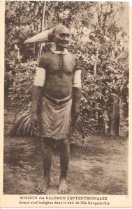 « Mission des Salomon Septentrionales. Grand chef indigène dans le sud de l’île Bougainville ». 13,8 x 8,8 cm. La coiffure de prestige en fibres que porte ce vieil homme n’est pas sans évoquer un certain nombre de figures en bois provenant des îles Salomon orientales présentant leurs cheveux tirés en arrière pour former une longue coiffure se terminant également en pointe. Réminiscence du bonnet d’initié upi de la société ruk-ruk, originaire des île Bougainville et Buka que les jeunes hommes portent durant leur parcours initiatique les conduisant à l’âge adulte. Ces bras sont lourdement ornés d’anneaux fa’ikome en tridacne ‘ima qui jouaient également un rôle important en tant que monnaie d’échange.