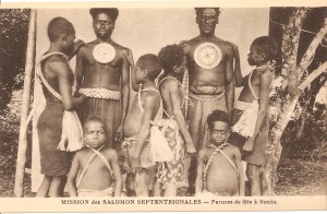 « Mission des Salomon Septentrionales — Parures de fête à Bouka ». 8,8 x 13,8 cm. Les deux hommes portent, sur la poitrine, un kapkap constitué d’un disque en coquille de bénitier recouvert d’un morceau d’écaille de tortue finement ajouré de motifs géométriques. Cette magnifique parure peut atteindre plus de vingt centimètres de diamètre.